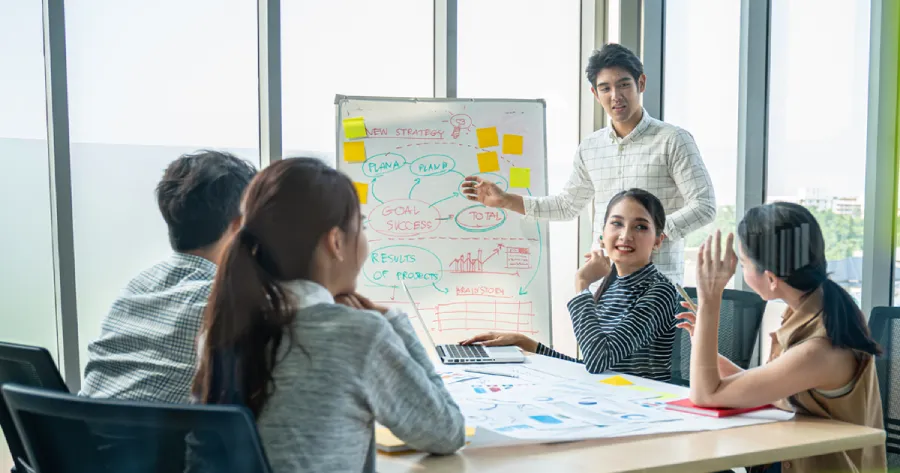 A team of 5 office coworkers are discussing, sitting around a table. One of them (a man) is standing next to a whiteboard pointing to the material written on it.