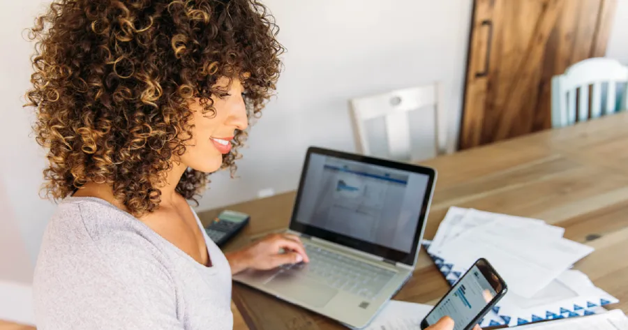 A woman is looking at her phone in her right hand and is working on a laptop with her left hand.