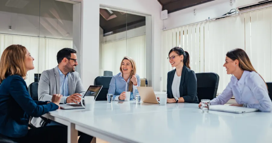 4 women and 1 man in professional attire are sitting around a table with their laptops.