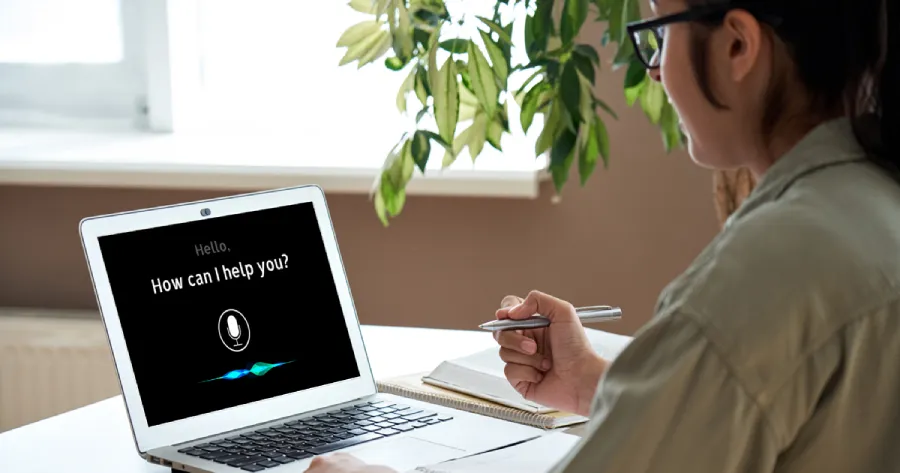 A women working on a laptop. The Laptop screen has the text How can I help You? With a microphone icon.