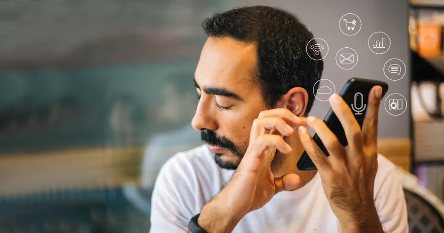 A man with a mobile phone near his ear. The mobile phone has small icons of a microphone, email, text box, shopping cart, wifi, cloud drive, etc, around it.