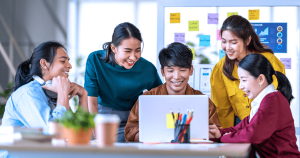  A group of 5 students sitting around a table working on a laptop.