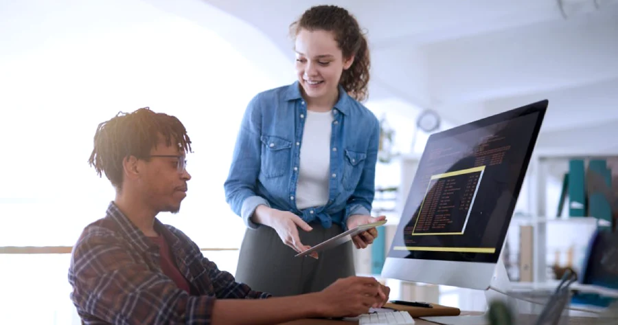 A man is working on a desktop. A woman is standing next to him showing him something on her tablet. 