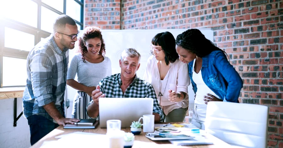 A group of 2 men and 3 women are looking at a laptop. One man out of the group is sitting on a chair in front of the laptop while the rest are standing around him. 