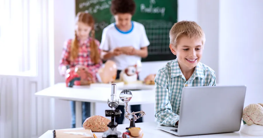 A boy with blond hair is studying on a laptop in a classroom.