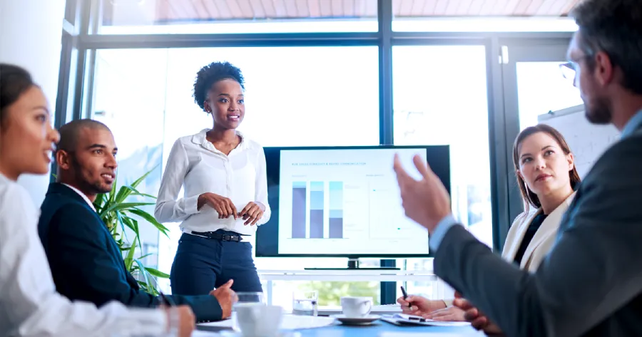 A team is sitting in a conference room and a woman is standing and showing a presentation. 