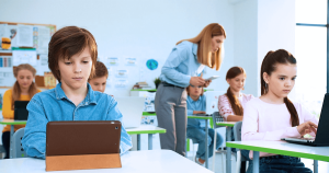 Students studying on digital devices in a classroom and a teacher is helping a student in the back of the classroom.