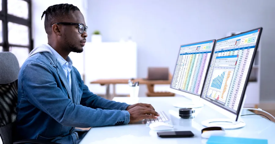 A man sitting at a desk has two desktop computer screens in front of him.