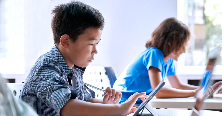 An Asian school boy is looking at a tablet with focus in a classroom.