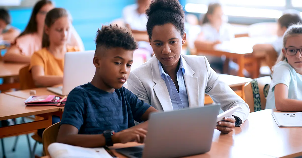  A Teacher and a student are sitting in a classroom studying from a laptop. 