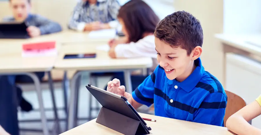 A little schoolboy, fully engaged with gamification in education, uses a tablet PC in a classroom.