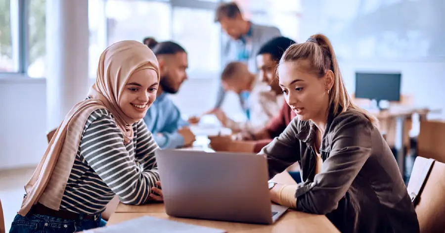 Two female students sitting opposite each other at a table in a college classroom, engrossed in a discussion about AI in higher education, while using a laptop.