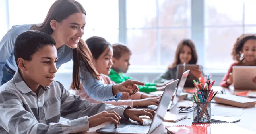 Teacher utilizing AI in K-12 education, teaches a school kid using a laptop in a classroom.