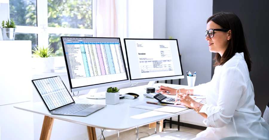 A woman sitting on a desk with two desktops is looking at the web accessibility audit report on a laptop on her desk. 