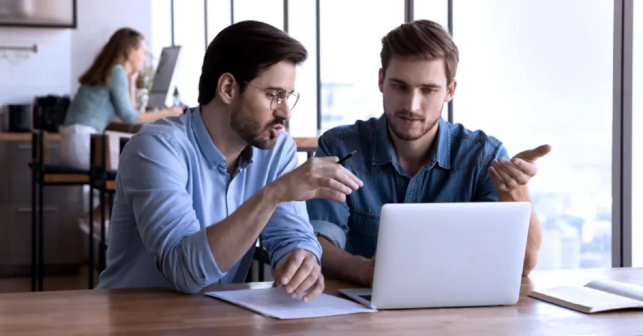 Two office employees engaged in workforce development, discussing a project on a laptop at a table. 