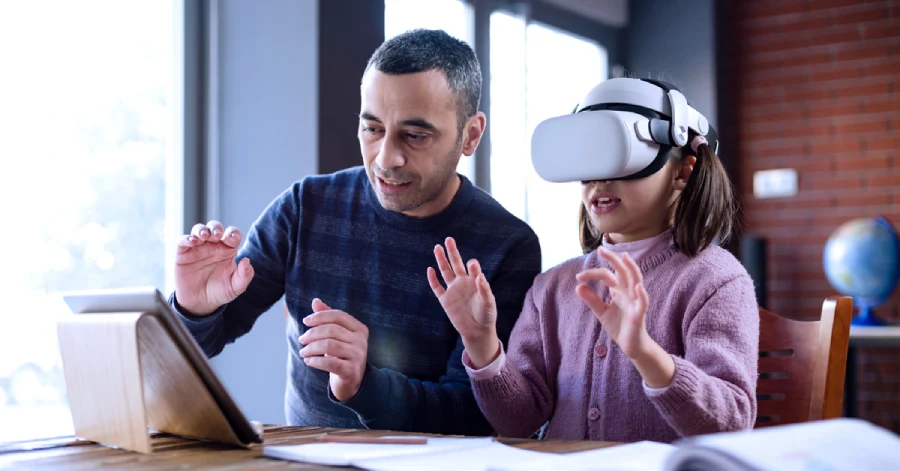 A teacher and a student are sitting together at a desk with a tablet. The student is fully immersed in a immersive learning program, wearing a VR headset.