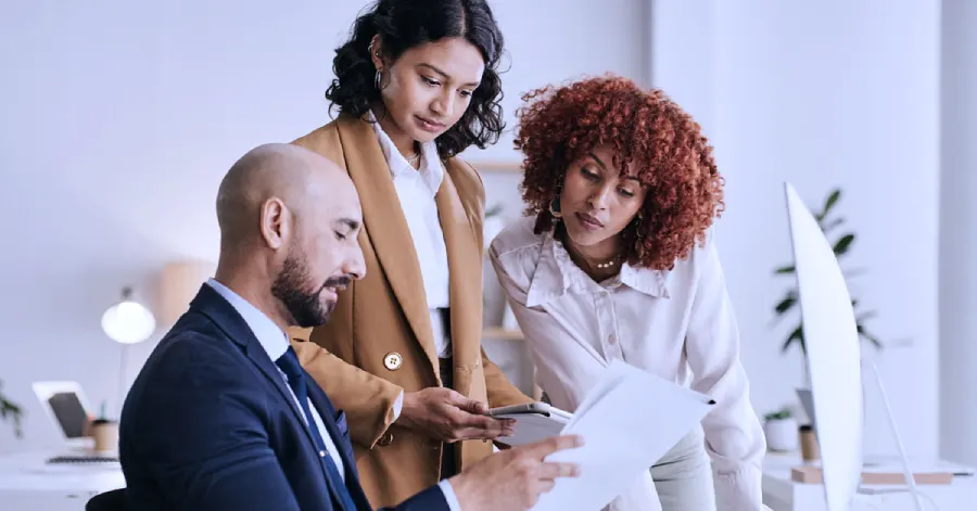 Three colleagues, two women and a man, huddle around a desk in an office, reviewing a document that outlines voluntary product accessibility guidelines. 