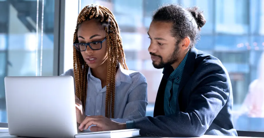 Concentrated diverse multiracial colleagues sit at a desk in an office, looking at a laptop screen and discussing a web accessibility audit.
