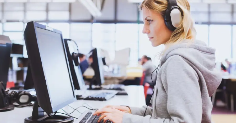 A woman wearing headphones using a desktop computer focused on working on a web accessibility audit at her desk in a busy office environment.